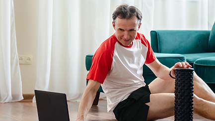 Person sitting on the floor with yoga/stretching equipment, laptop positioned in front of them to follow along during a workshop. 