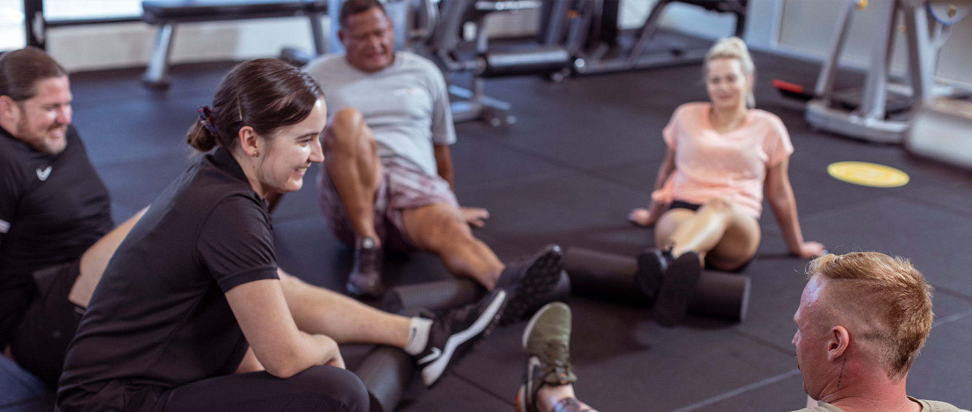 Group in a gym space, sitting on the floor stretching and rolling muscles with a Mates4Mates exercise physiologist.
