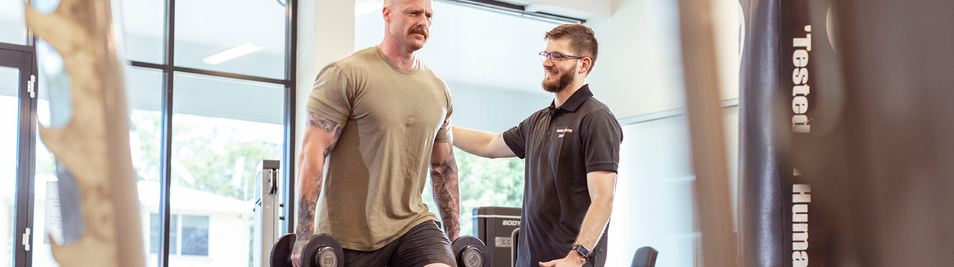 Exercise physiologist supporting a client in the gym as they perform an exercise with dumbbell weights.